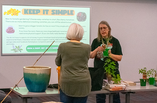 Woman presents a gardening slide titled KEEP IT SIMPLE while another woman listens beside a table of potted plants.